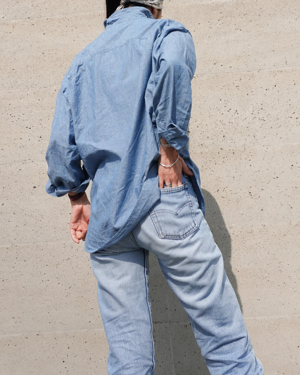 Chemise en chambray bleu Merci Paris en coton, portée avec un jean, vue de dos sur un mur en béton texturé
