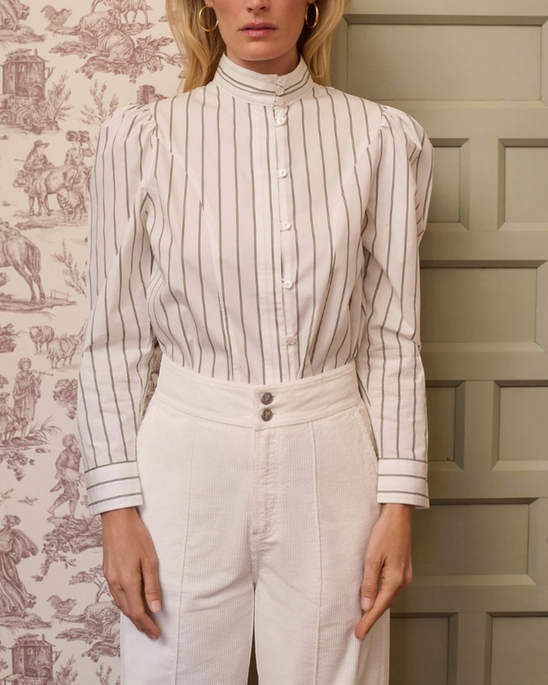 Femme portant la chemise Iza Merci Paris à rayures kaki, col mao et épaules bouffantes devant un papier peint toile